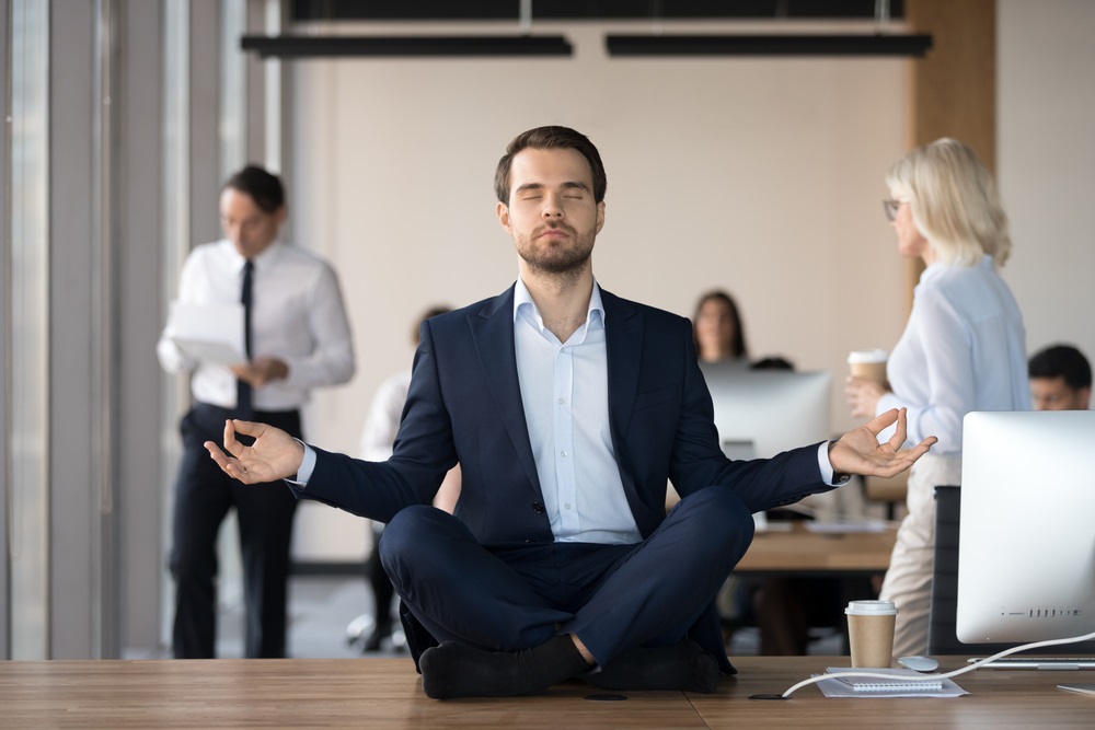 Mindful calm businessman in suit meditating at office sitting in lotus position on work desk, successful ceo executive doing yoga exercise at workplace, peace of mind, no stress free relief concept